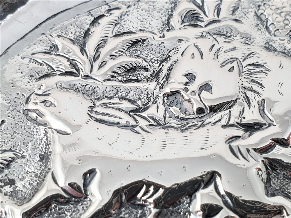 An oval silver dish featuring a chased repousse hunting scene in relief with a leaf pattern border. View IX
