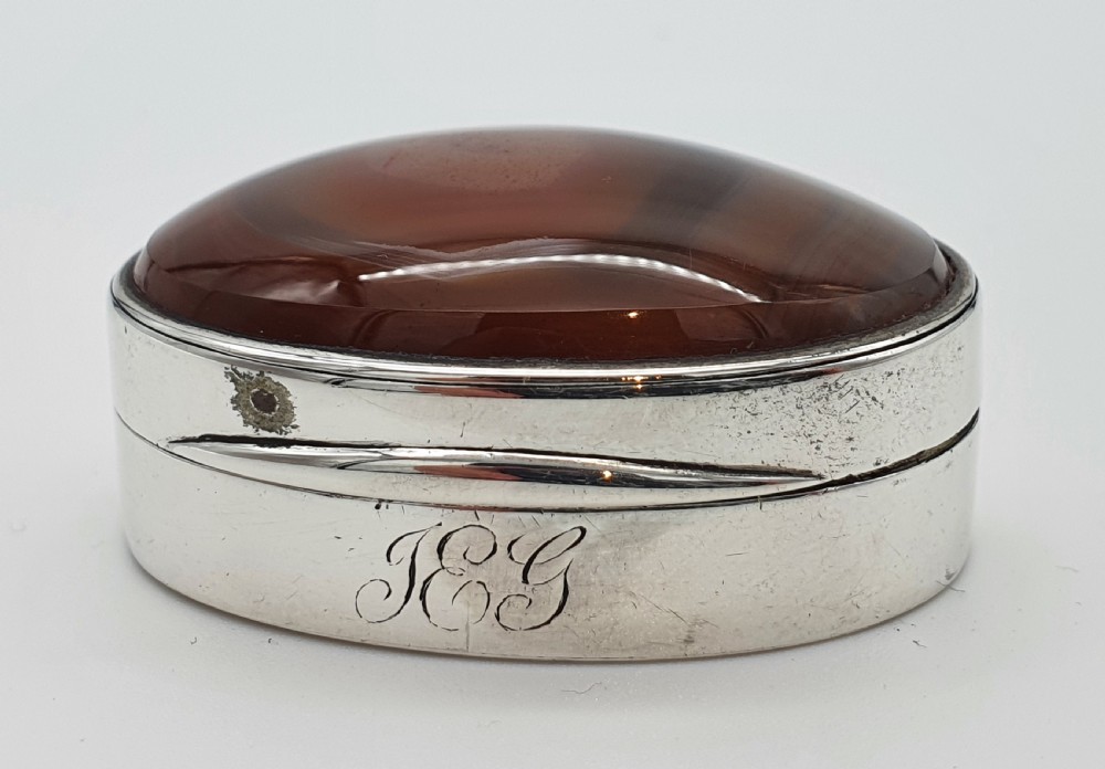 A Georgian oval silver vinaigrette featuring a smooth brown agate top and engraved script monogram on the silver collar.