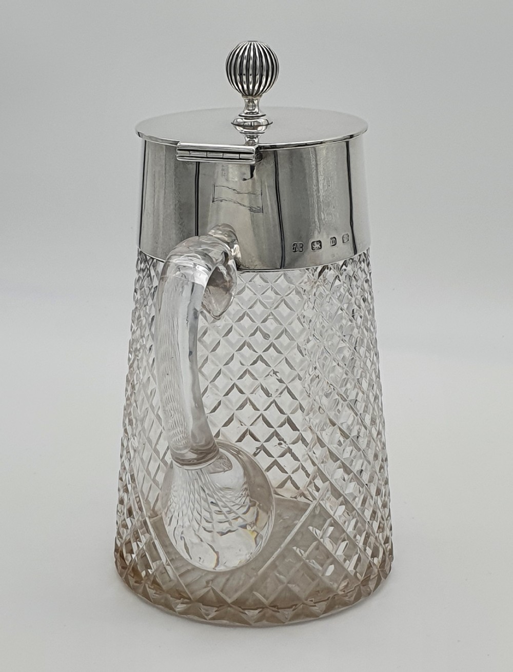 A late Victorian claret jug featuring a diamond-cut clear glass body paired with a polished sterling silver collar, hinged flat lid, fluted finial, and clear glass handle. View II