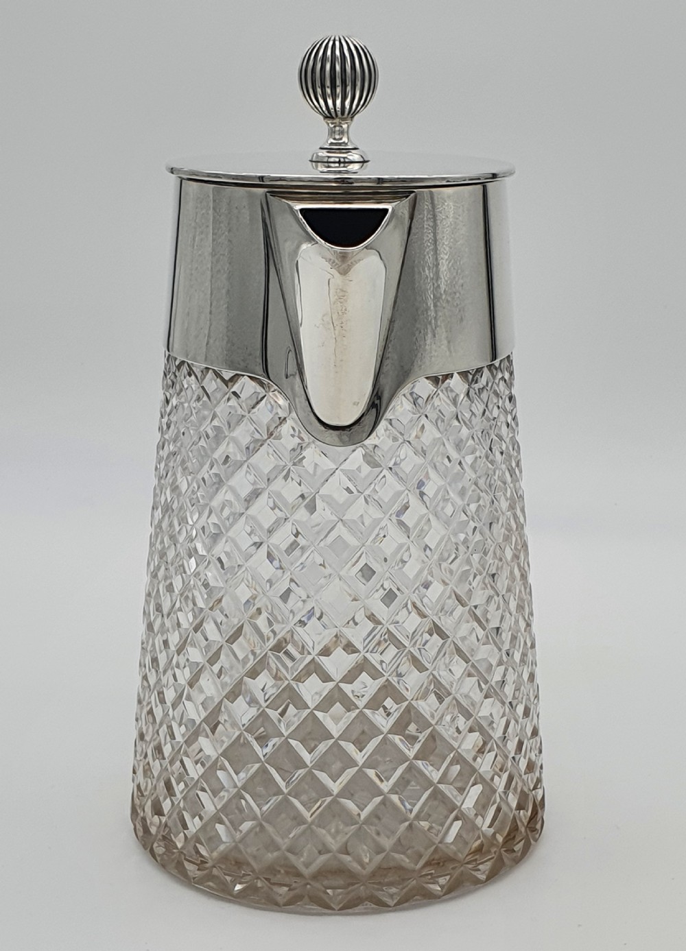 A late Victorian claret jug featuring a diamond-cut clear glass body paired with a polished sterling silver collar, hinged flat lid, fluted finial, and clear glass handle. View V