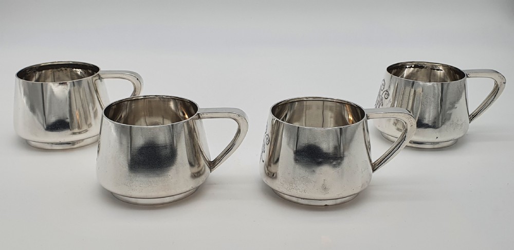 A set of four small silver vodka cups with angular handles and engraved monograms against a white backdrop.