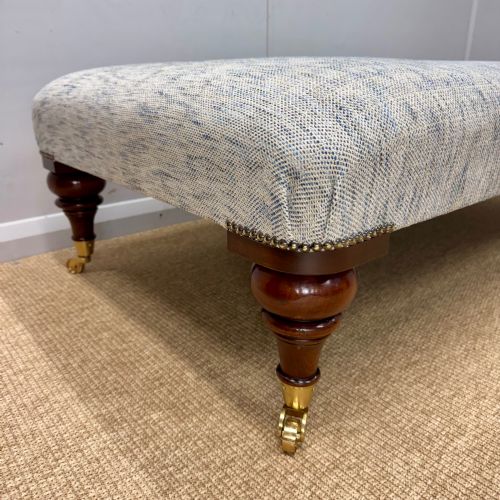 fig 1. A low rectangular upholstered footstool featuring light blue and grey woven fabric, decorative brass tack trim, and turned dark mahogany legs on brass castors.