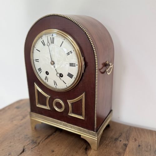 fig 1. An arched antique French mahogany mantel clock featuring a white face with Roman numerals, decorative brass trim, and a stepped brass base.