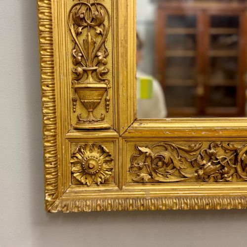 fig 1. A large horizontal rectangular mirror featuring an intricately carved giltwood frame with classical urn, floral, and leaf motifs.