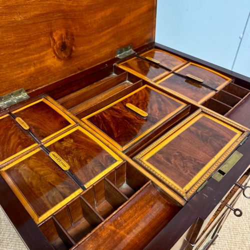 fig 1. A late Georgian mahogany work table with an open hinged lid revealing fitted interior compartments, pleated fabric side panels, and square tapered legs on brass castors.
