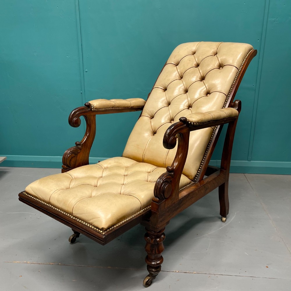 A Late Regency mahogany reclining armchair featuring buttoned tan leather upholstery, brass stud trim, scroll arms, and turned front legs on castors. View II