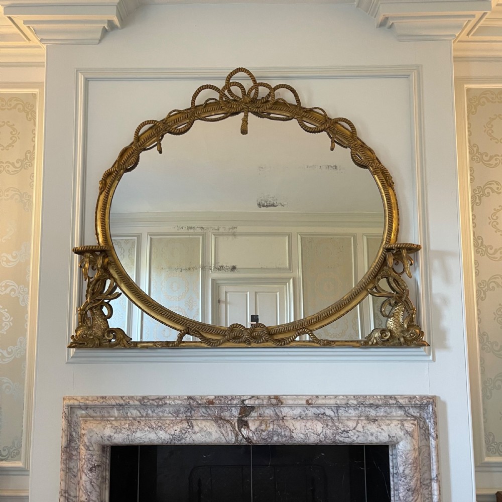 A large French gilt overmantel mirror featuring an oval glass plate surrounded by rope twist mouldings, a ribbon crest, and carved dolphin corner supports. View II