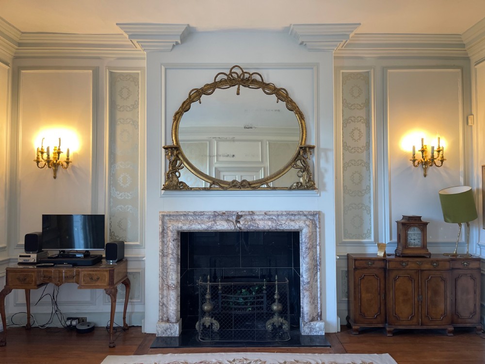 A large French gilt overmantel mirror featuring an oval glass plate surrounded by rope twist mouldings, a ribbon crest, and carved dolphin corner supports. View V