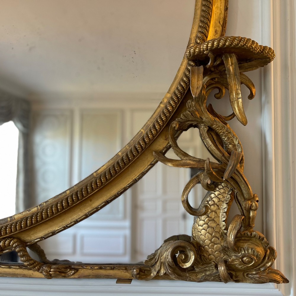 A large French gilt overmantel mirror featuring an oval glass plate surrounded by rope twist mouldings, a ribbon crest, and carved dolphin corner supports. View VI