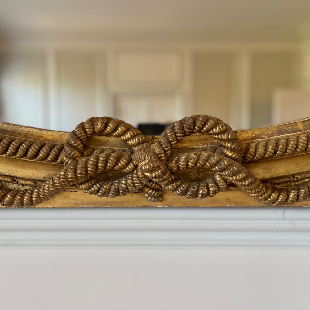 A large French gilt overmantel mirror featuring an oval glass plate surrounded by rope twist mouldings, a ribbon crest, and carved dolphin corner supports. View VII