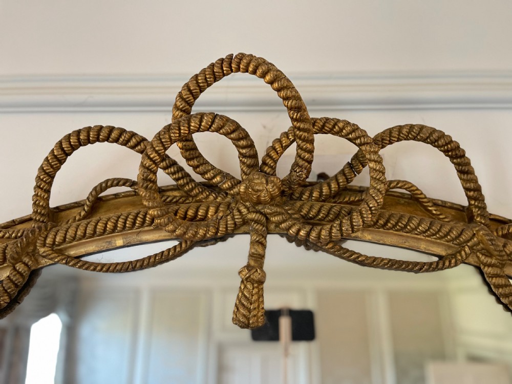 A large French gilt overmantel mirror featuring an oval glass plate surrounded by rope twist mouldings, a ribbon crest, and carved dolphin corner supports. View VIII