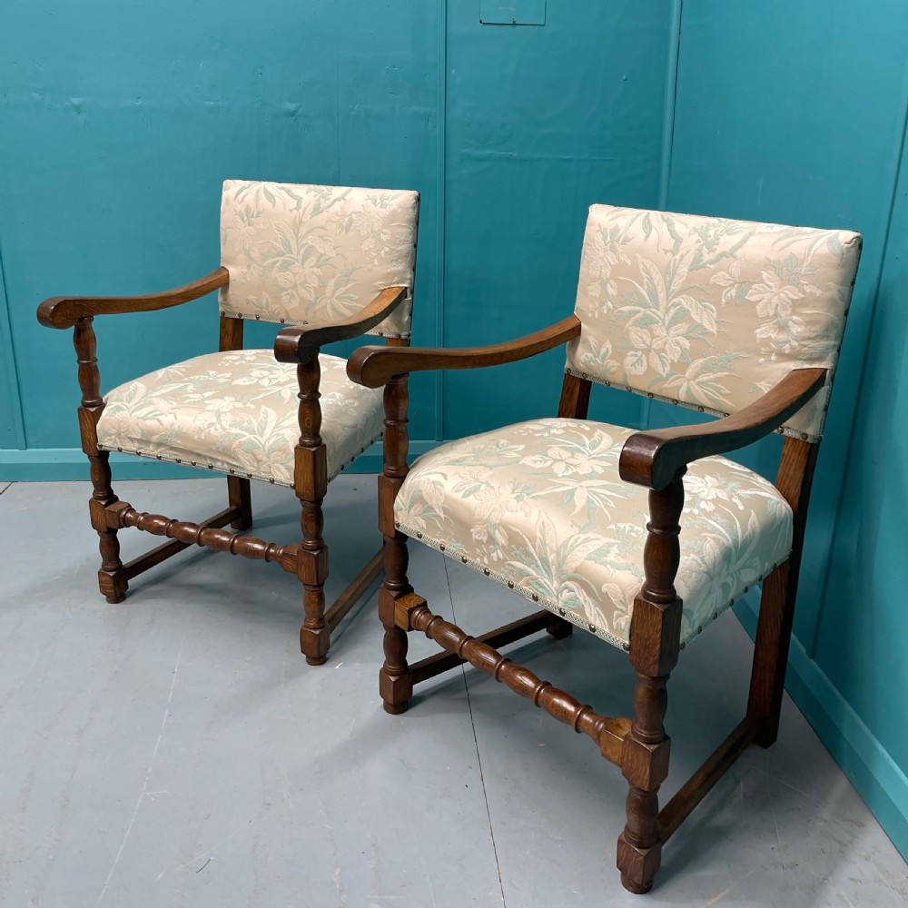 A pair of English oak armchairs featuring turned legs, stretchers, open curved armrests, and floral pattern cream upholstery secured with metal studs.