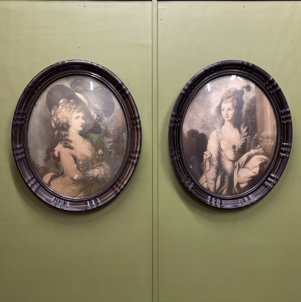 A pair of matching oval portraits featuring 18th-century style figures, housed under convex glass within dark moulded frames.