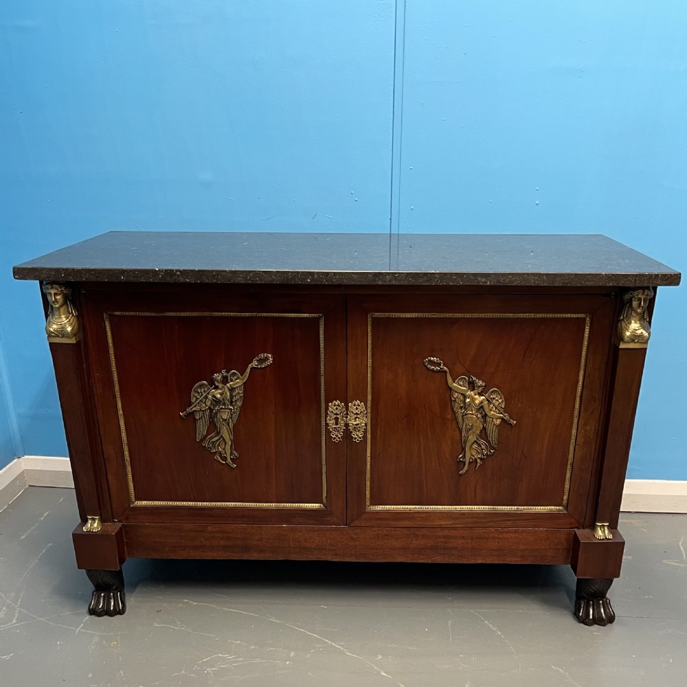 A French Empire mahogany commode a portes featuring a dark marble top, gilt bronze mounts of winged figures, and carved paw feet.