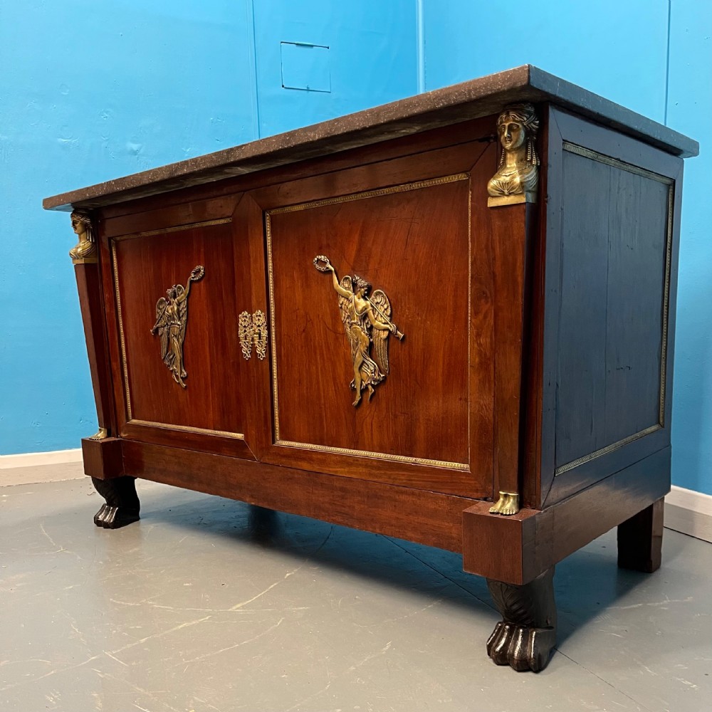 A French Empire mahogany commode a portes featuring a dark marble top, gilt bronze mounts of winged figures, and carved paw feet. View II