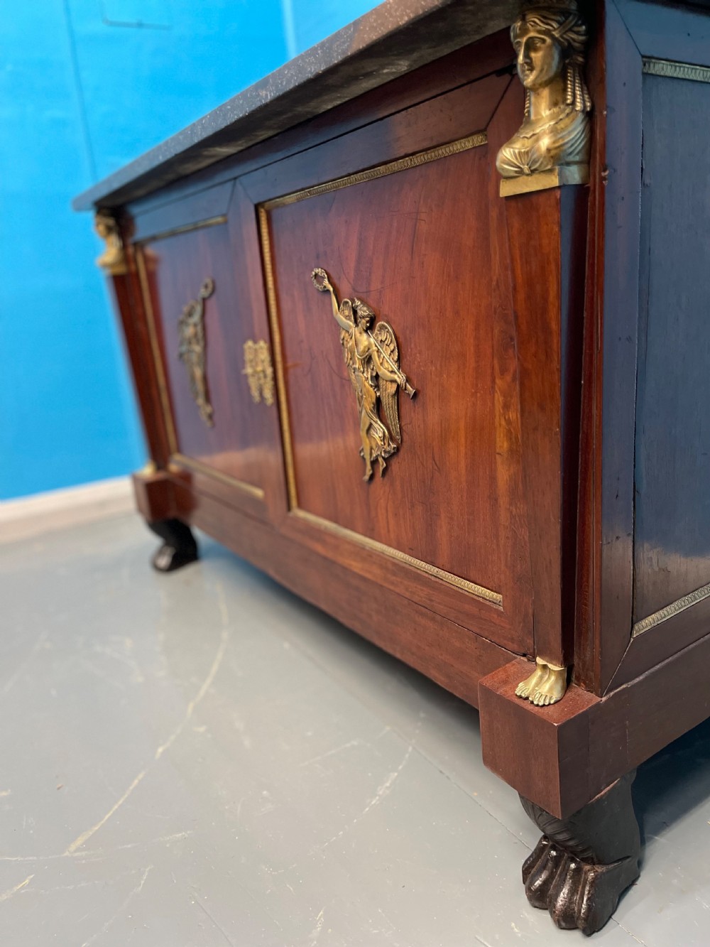 A French Empire mahogany commode a portes featuring a dark marble top, gilt bronze mounts of winged figures, and carved paw feet. View III