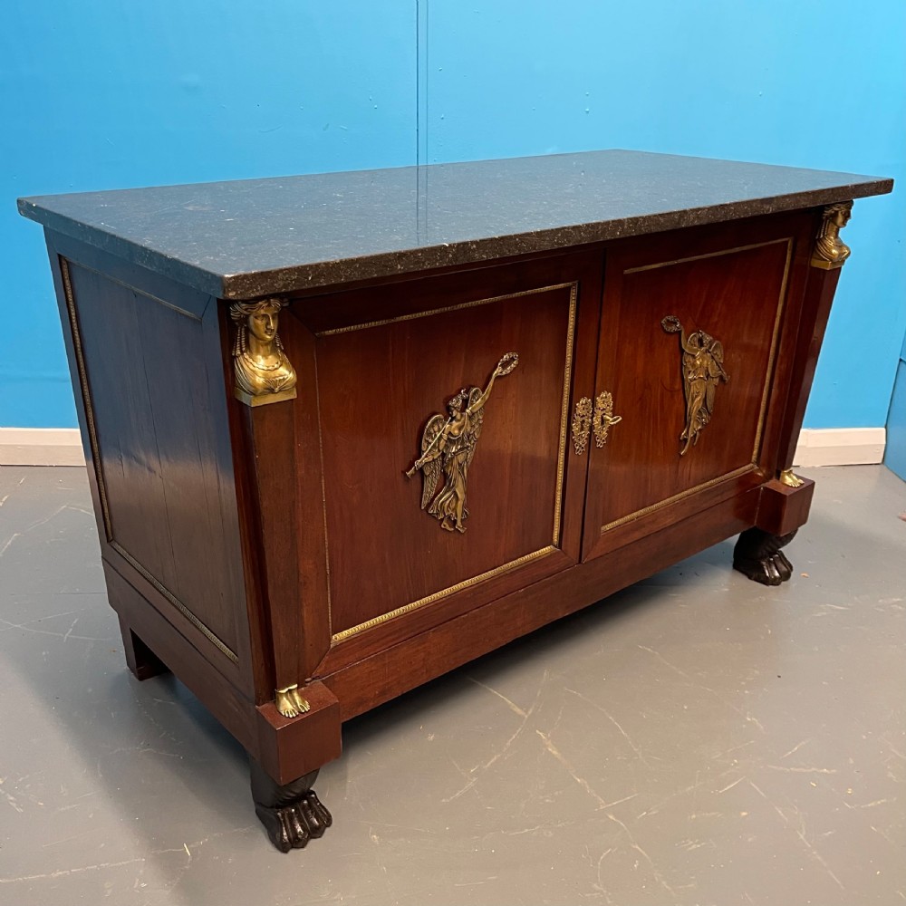 A French Empire mahogany commode a portes featuring a dark marble top, gilt bronze mounts of winged figures, and carved paw feet. View V