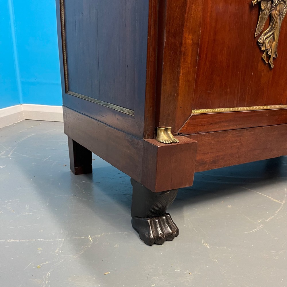A French Empire mahogany commode a portes featuring a dark marble top, gilt bronze mounts of winged figures, and carved paw feet. View VII