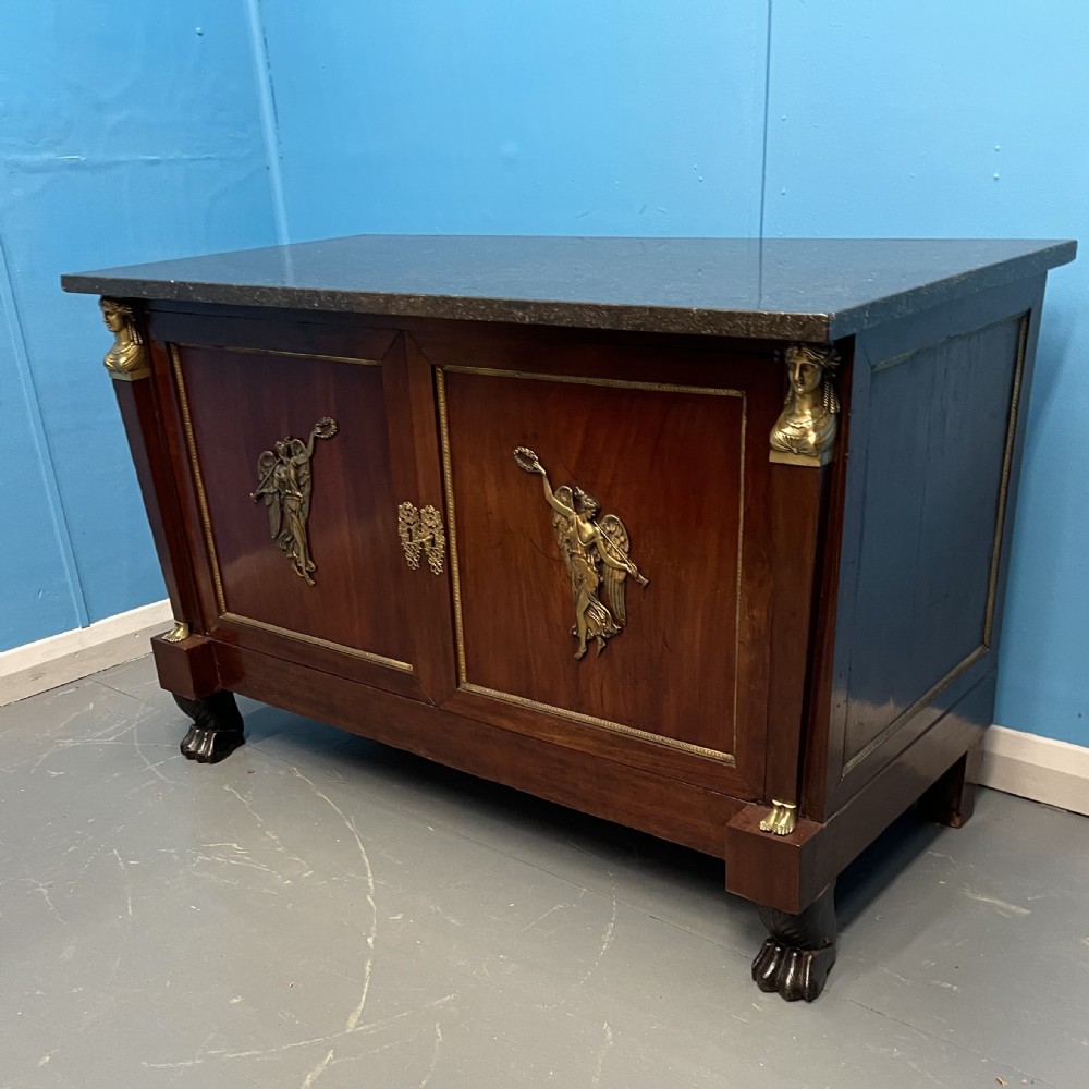 A French Empire mahogany commode a portes featuring a dark marble top, gilt bronze mounts of winged figures, and carved paw feet. View VIII