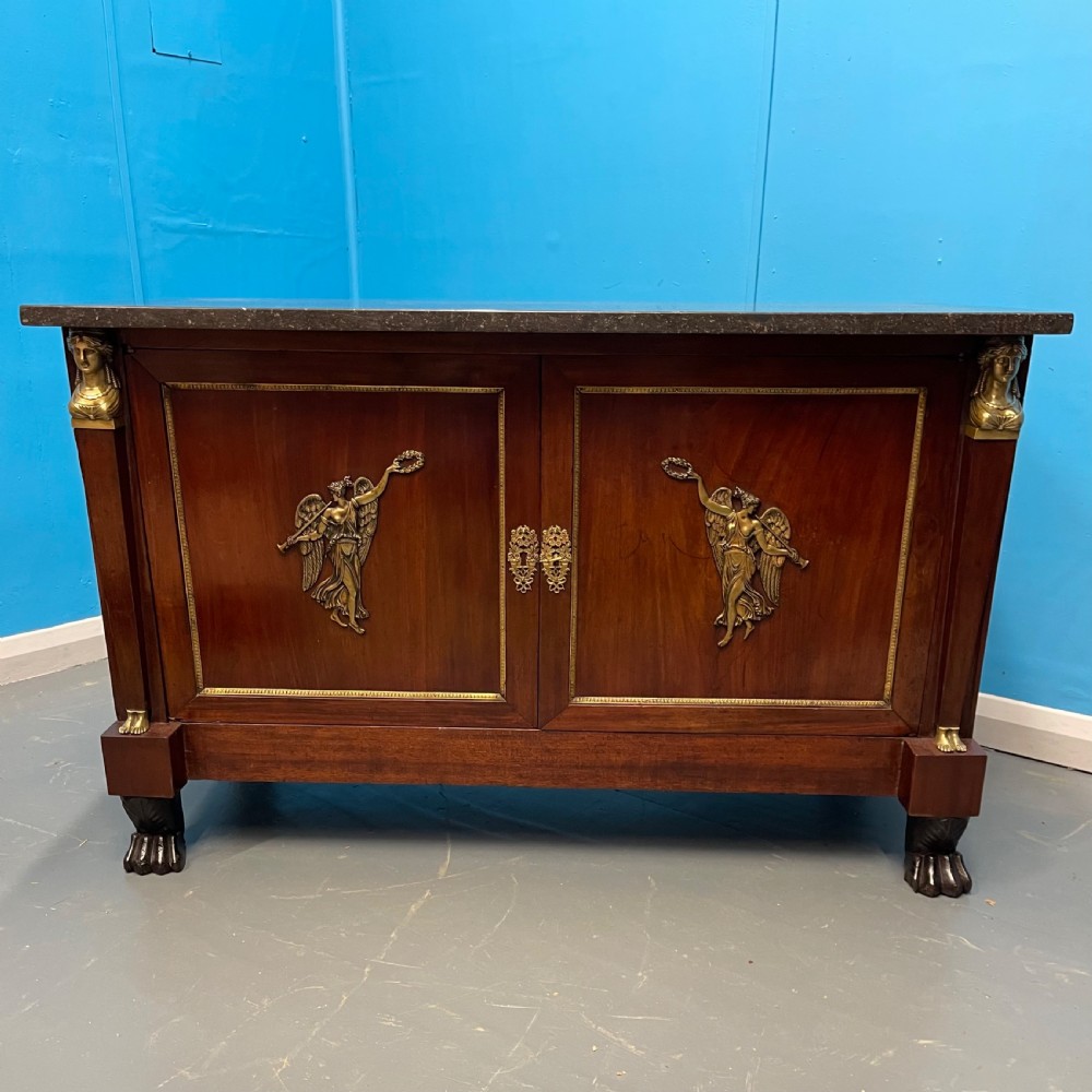 A French Empire mahogany commode a portes featuring a dark marble top, gilt bronze mounts of winged figures, and carved paw feet. View IX