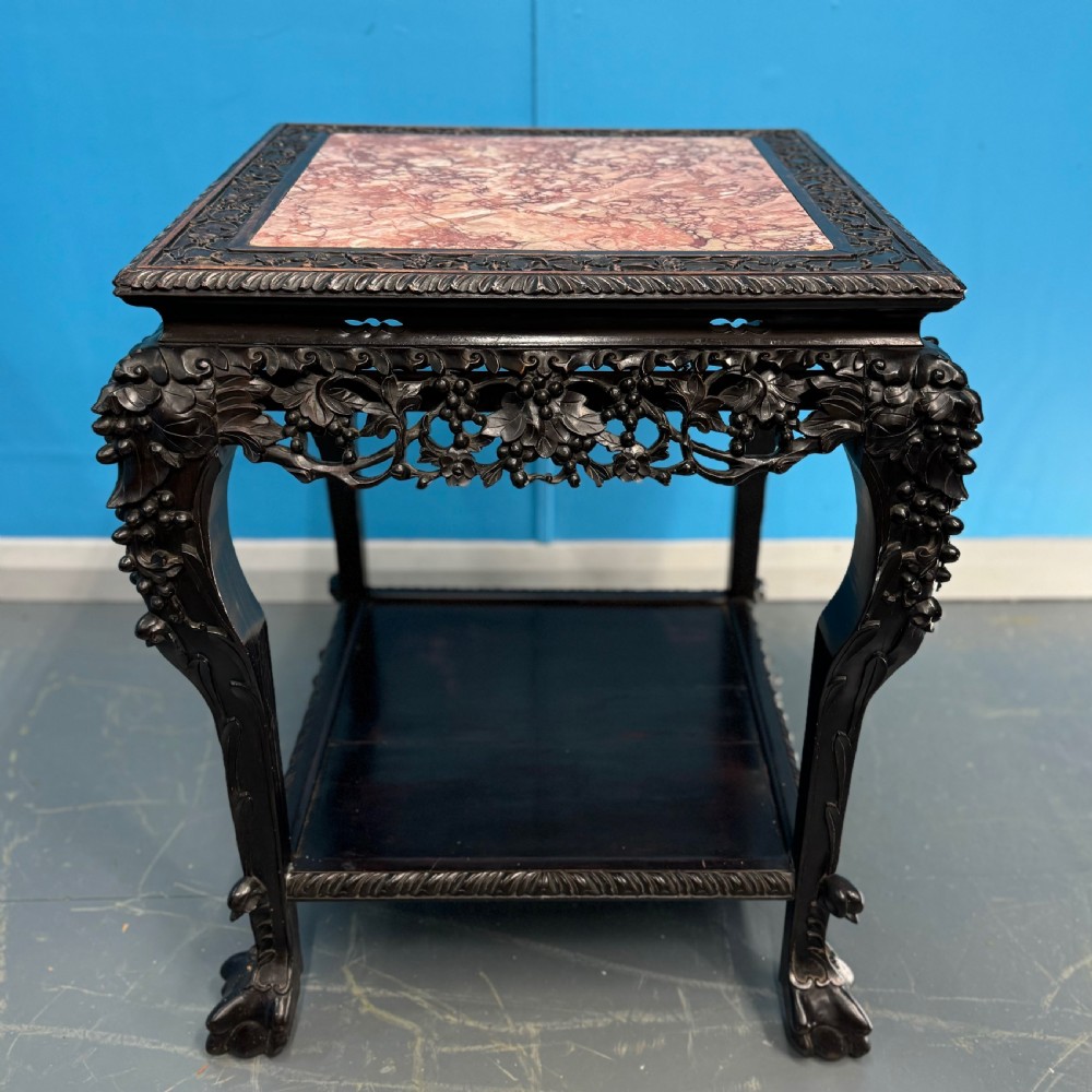 A dark carved Chinese hardwood side table with an inset pink marble top, openwork grapevine apron, cabriole legs, and a lower shelf.