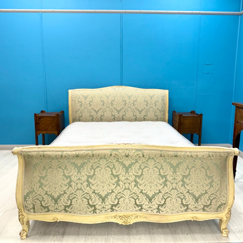 A king-size French bed frame featuring a carved cream-painted timber frame upholstered in green damask fabric with a scroll-end footboard.