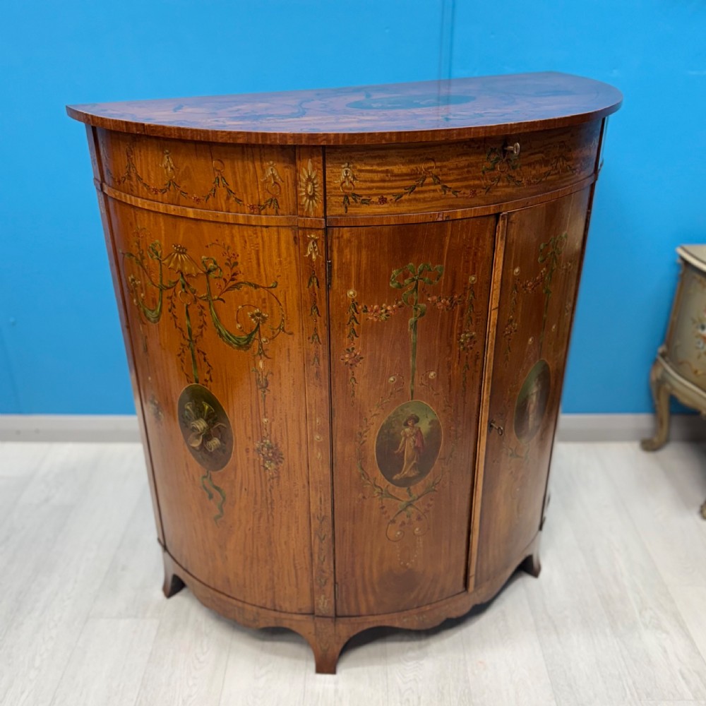 A 19th-century demi-lune satinwood side cabinet featuring hand-painted floral swags, ribbon motifs, and portrait medallions across its curved front doors.