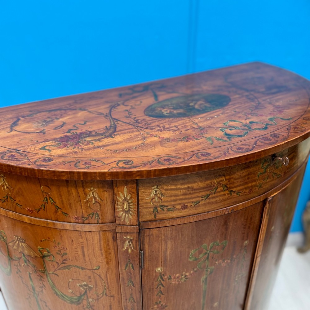 A 19th-century demi-lune satinwood side cabinet featuring hand-painted floral swags, ribbon motifs, and portrait medallions across its curved front doors. View V