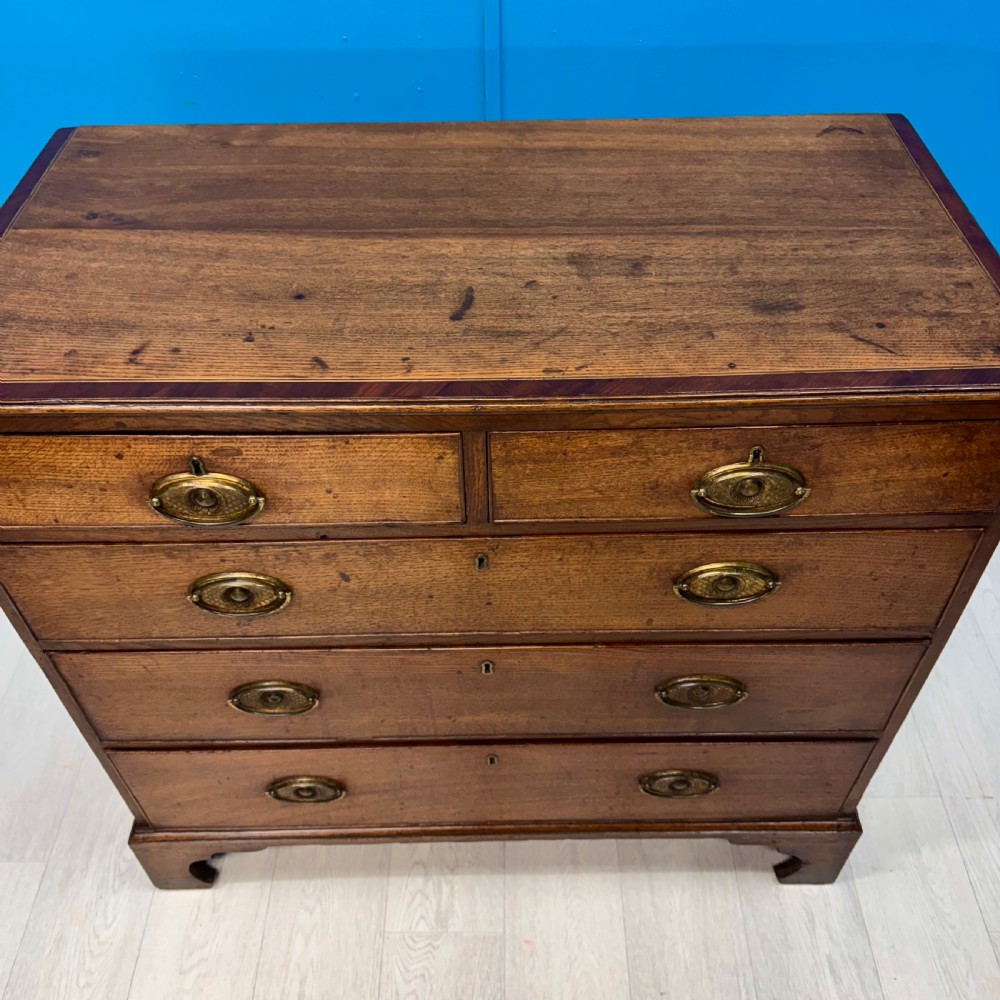 A Georgian oak chest of drawers featuring two short drawers over three long drawers with oval brass drop handles and bracket feet. View V