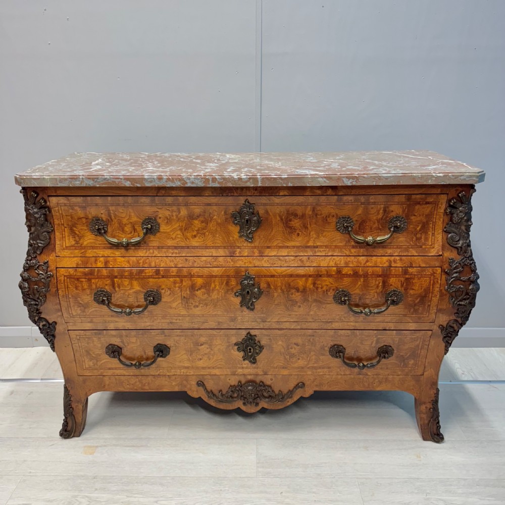 A French Louis XV style burr walnut commode featuring a curved bombee profile, three drawers with ornate brass handles, metal corner mounts, and a shaped marble top.