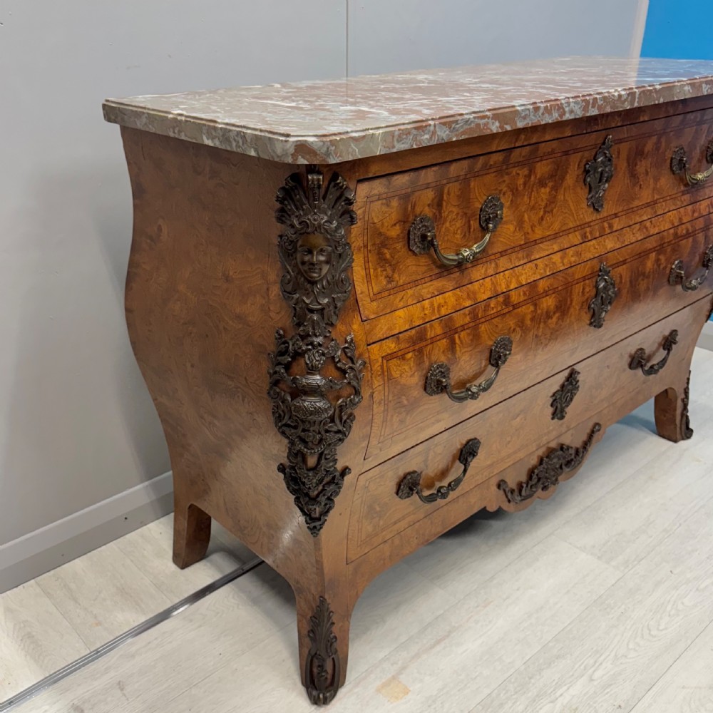 A French Louis XV style burr walnut commode featuring a curved bombee profile, three drawers with ornate brass handles, metal corner mounts, and a shaped marble top. View III