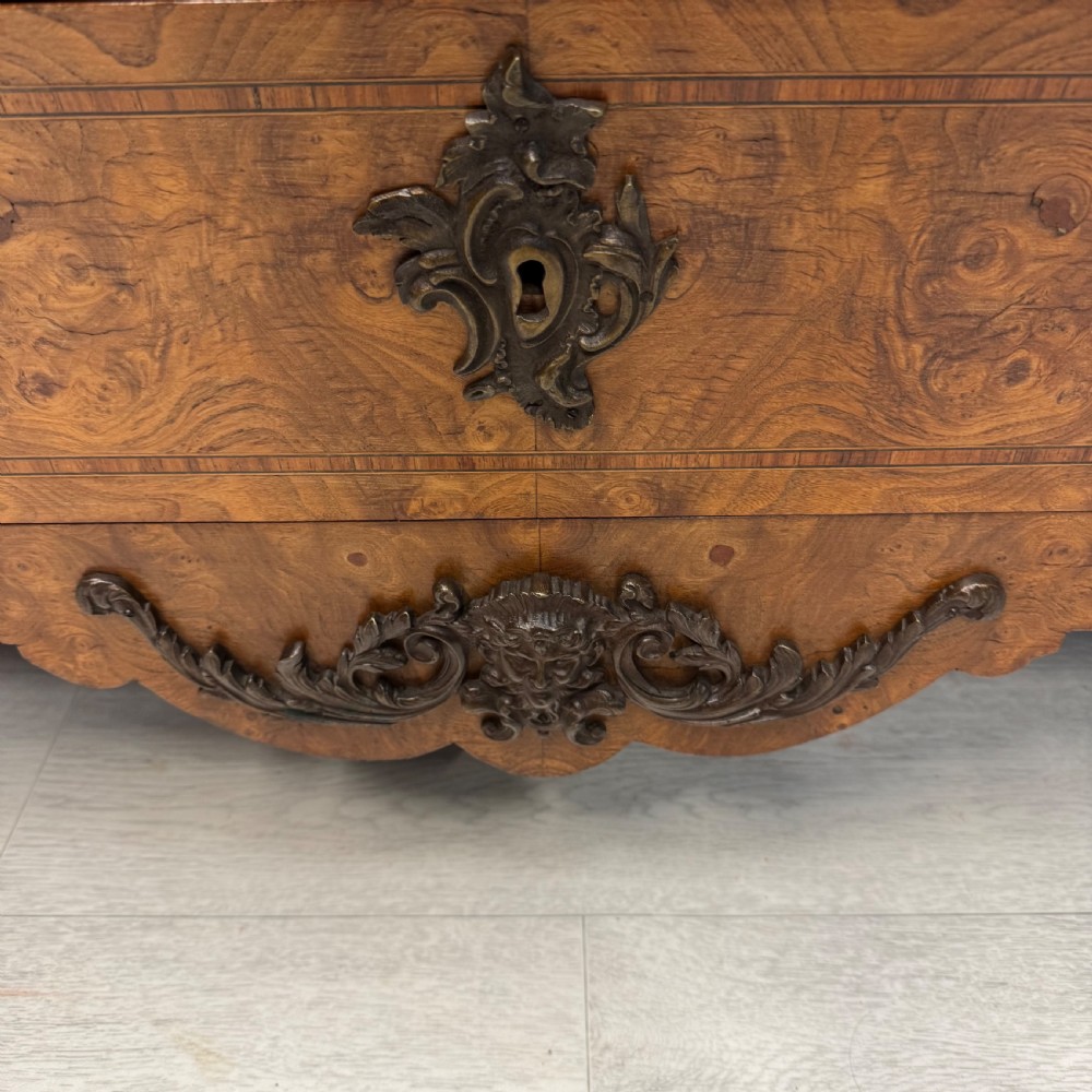 A French Louis XV style burr walnut commode featuring a curved bombee profile, three drawers with ornate brass handles, metal corner mounts, and a shaped marble top. View V