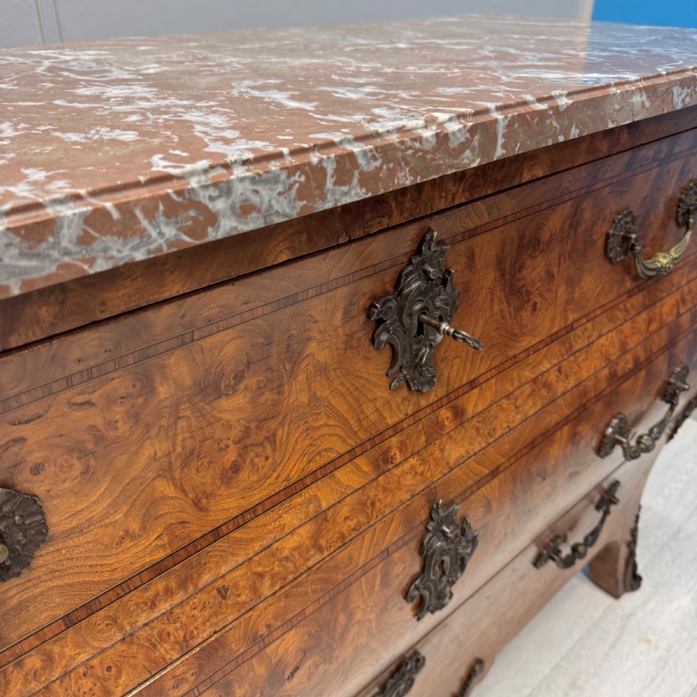 A French Louis XV style burr walnut commode featuring a curved bombee profile, three drawers with ornate brass handles, metal corner mounts, and a shaped marble top. View VI