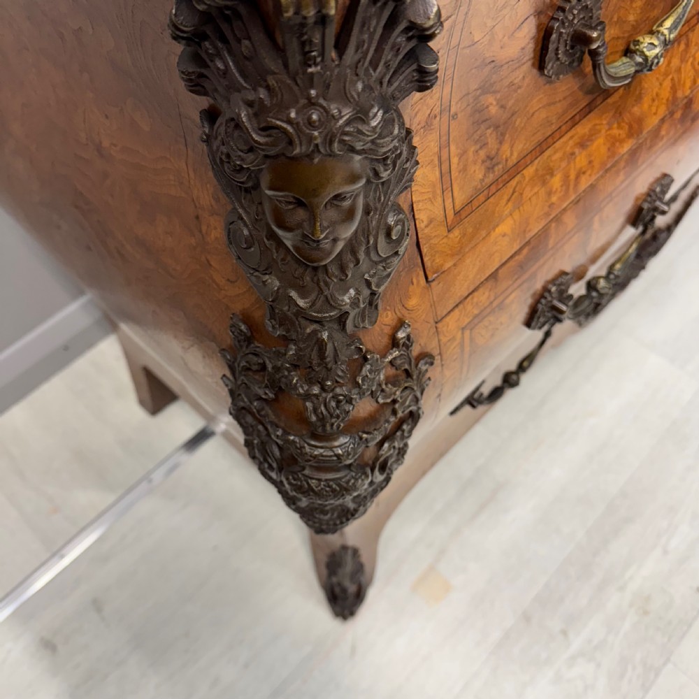 A French Louis XV style burr walnut commode featuring a curved bombee profile, three drawers with ornate brass handles, metal corner mounts, and a shaped marble top. View VII