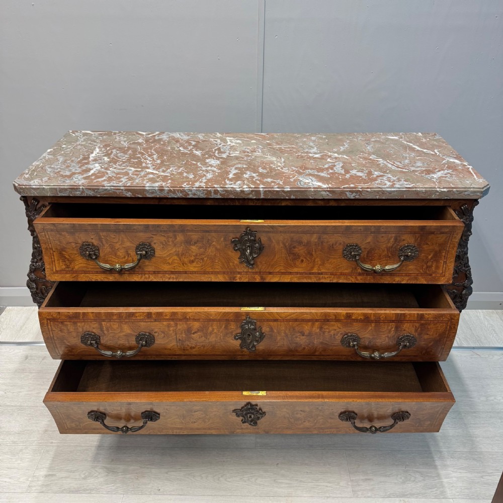 A French Louis XV style burr walnut commode featuring a curved bombee profile, three drawers with ornate brass handles, metal corner mounts, and a shaped marble top. View VIII