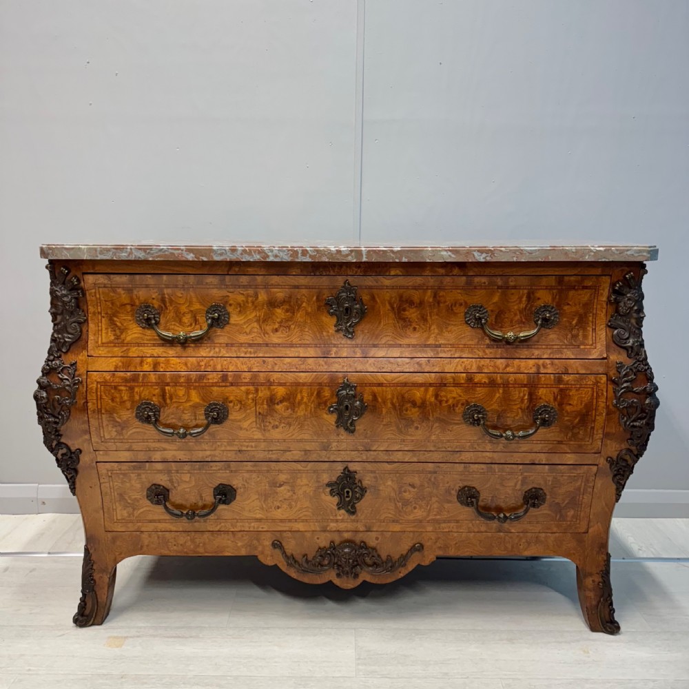 A French Louis XV style burr walnut commode featuring a curved bombee profile, three drawers with ornate brass handles, metal corner mounts, and a shaped marble top. View IX