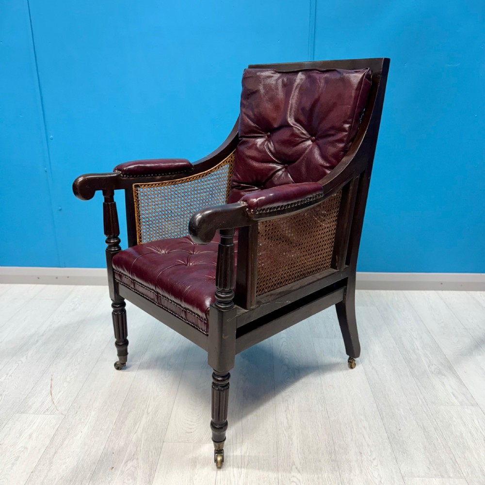 An early 19th-century mahogany bergere armchair with button-tufted burgundy leather cushions, woven cane side panels, turned front legs, and brass castors.