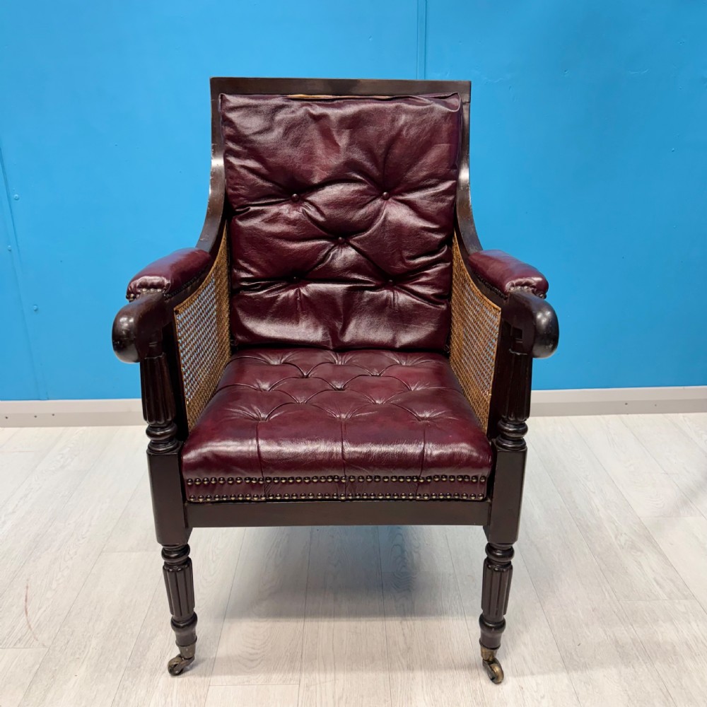 An early 19th-century mahogany bergere armchair with button-tufted burgundy leather cushions, woven cane side panels, turned front legs, and brass castors. View II