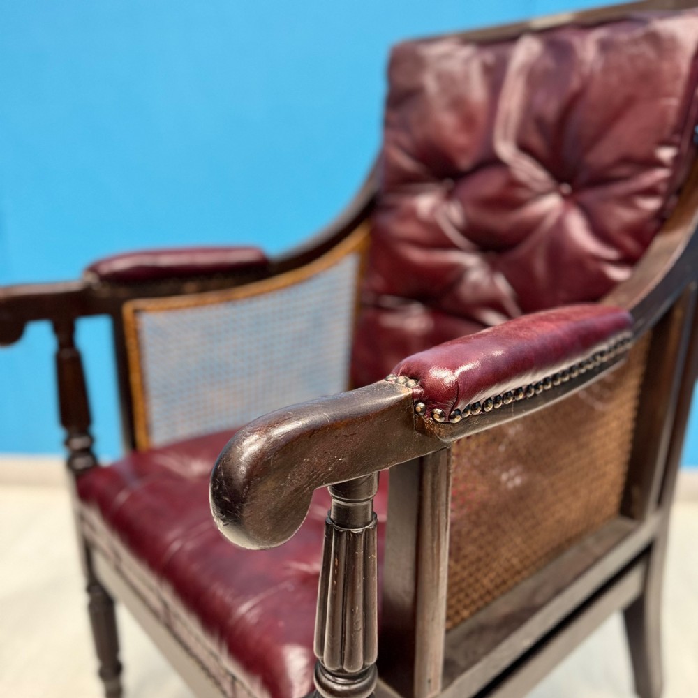 An early 19th-century mahogany bergere armchair with button-tufted burgundy leather cushions, woven cane side panels, turned front legs, and brass castors. View III
