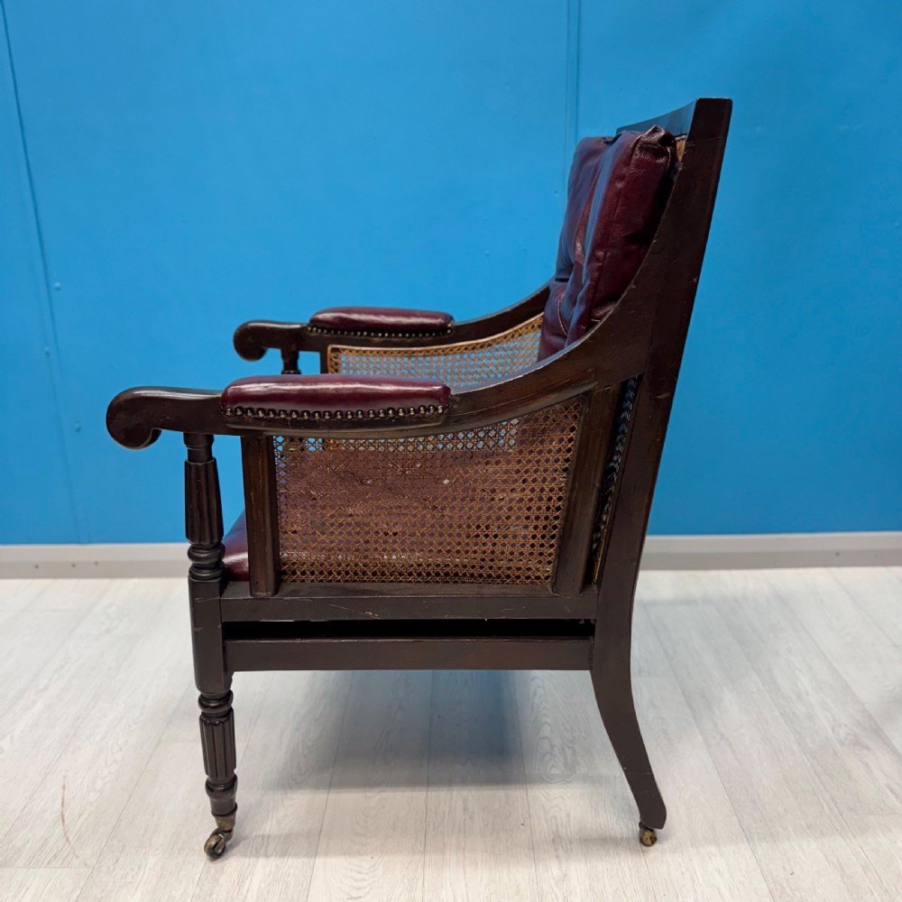 An early 19th-century mahogany bergere armchair with button-tufted burgundy leather cushions, woven cane side panels, turned front legs, and brass castors. View V