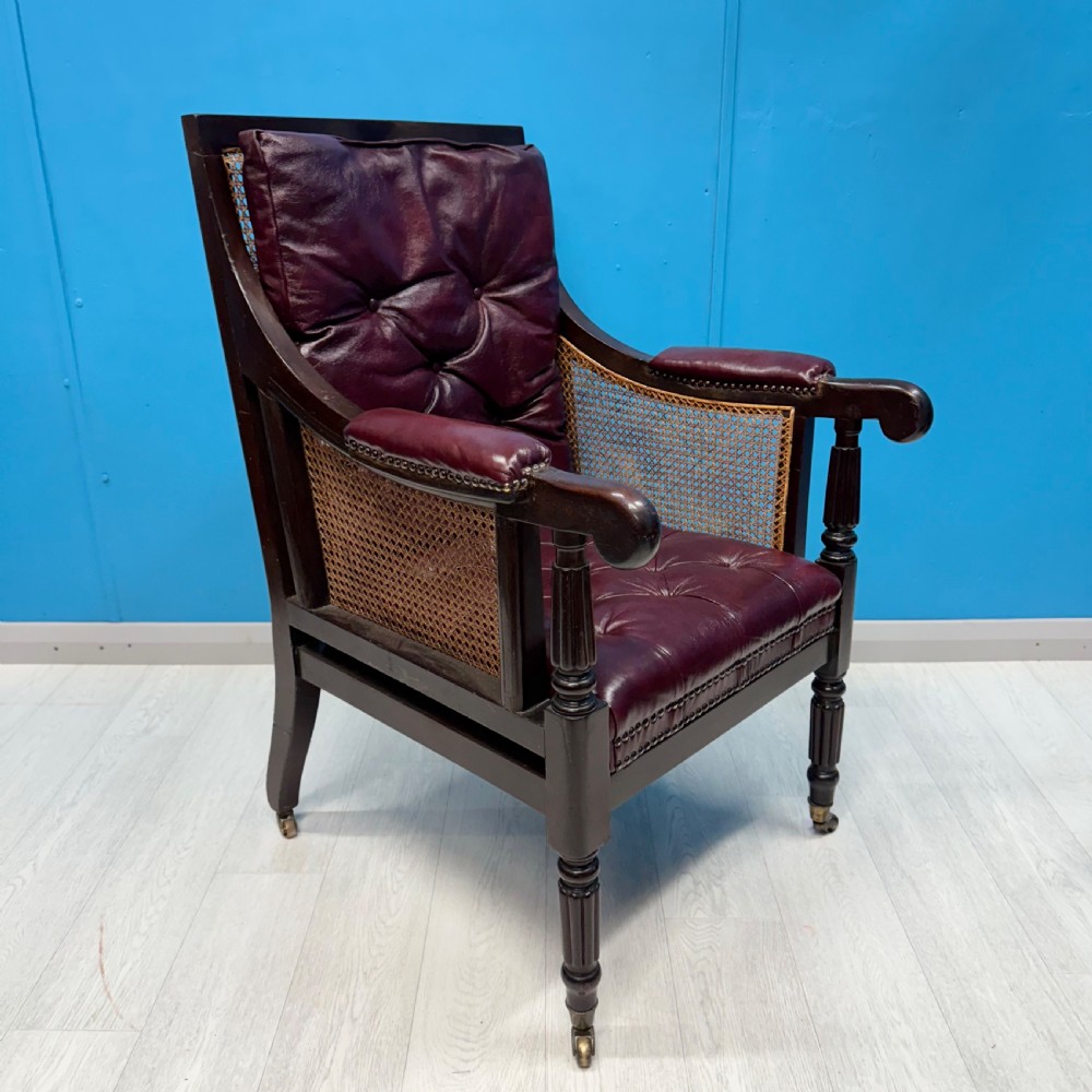 An early 19th-century mahogany bergere armchair with button-tufted burgundy leather cushions, woven cane side panels, turned front legs, and brass castors. View VI