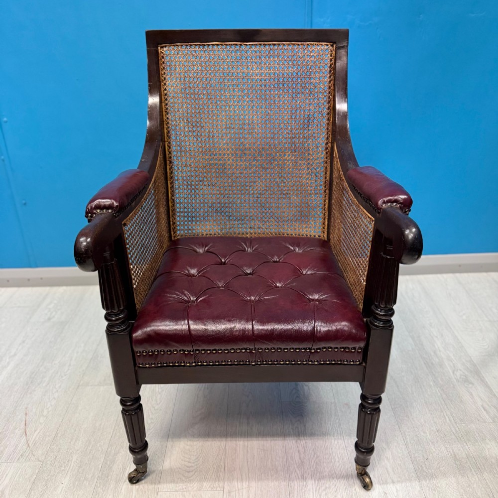 An early 19th-century mahogany bergere armchair with button-tufted burgundy leather cushions, woven cane side panels, turned front legs, and brass castors. View VII