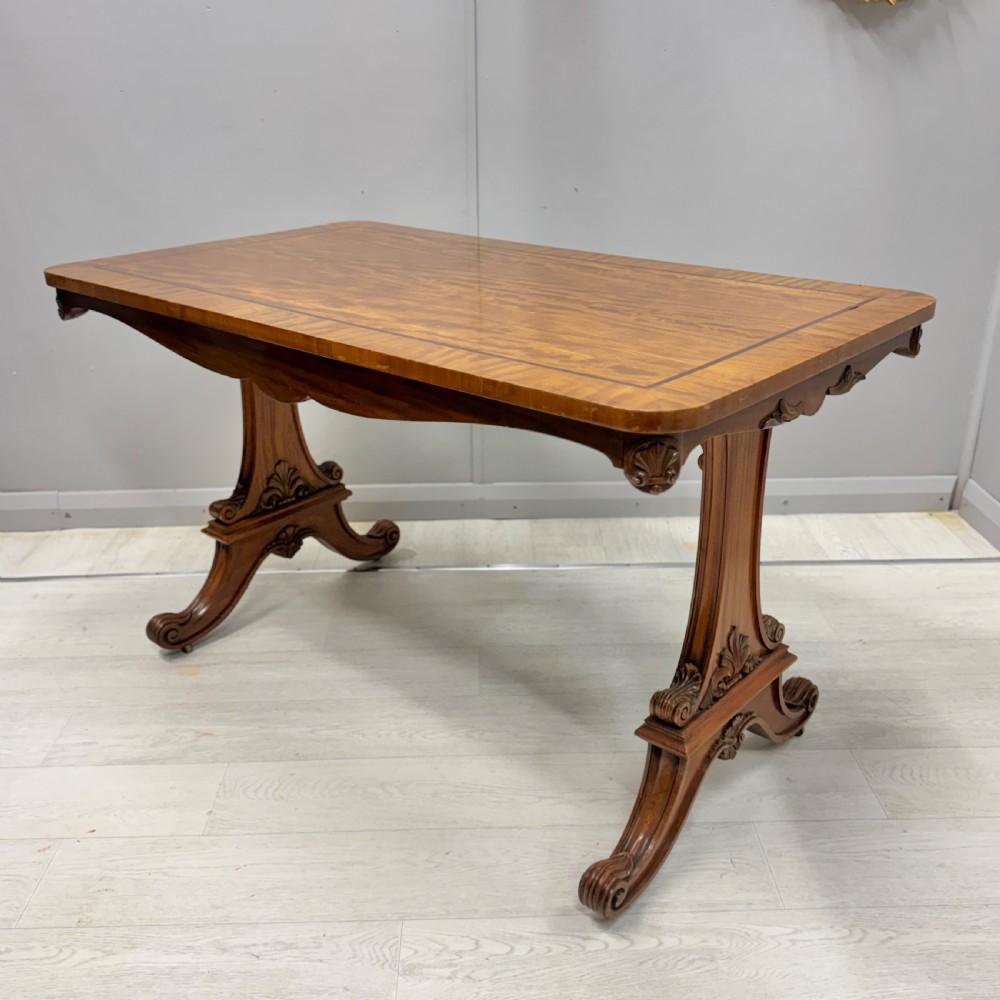 A 19th-century satinwood sofa table featuring a rectangular crossbanded top supported by carved trestle end supports and splayed legs on castors.
