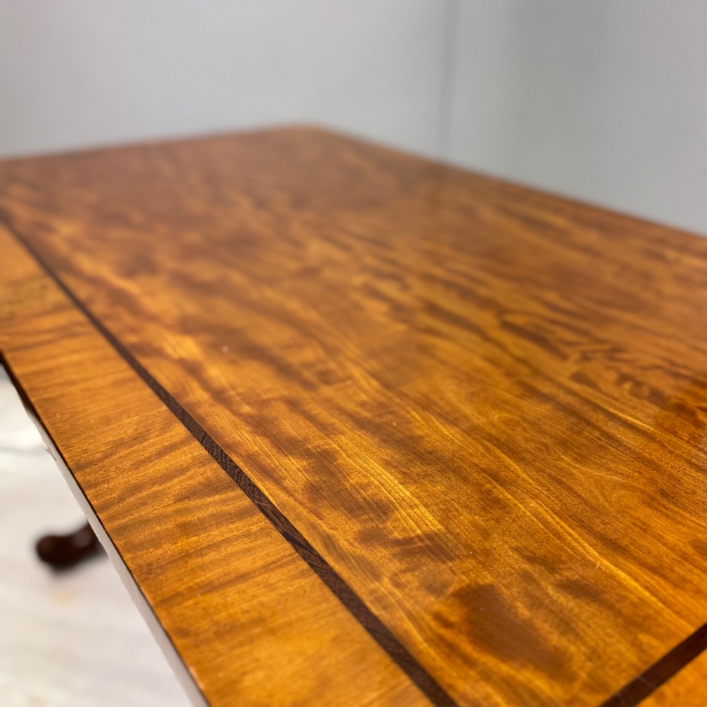 A 19th-century satinwood sofa table featuring a rectangular crossbanded top supported by carved trestle end supports and splayed legs on castors. View VI