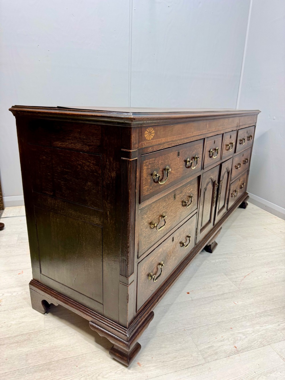 An 18th-century Georgian oak dresser base featuring inlaid fan motifs on the top frieze, multiple drawers with brass swan-neck handles, and twin central panelled cupboard doors. View VIII