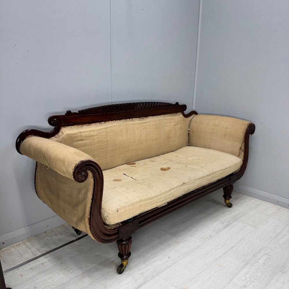 An antique Regency mahogany sofa frame featuring carved scroll arms, fluted turned legs on brass castors, and calico upholstery prepared for recovering. View III