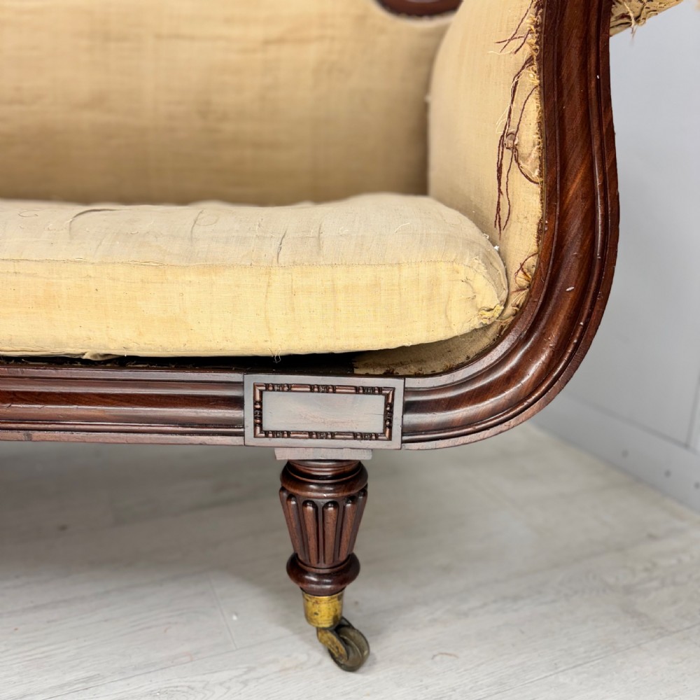 An antique Regency mahogany sofa frame featuring carved scroll arms, fluted turned legs on brass castors, and calico upholstery prepared for recovering. View VI