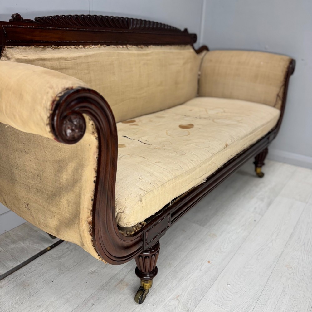 An antique Regency mahogany sofa frame featuring carved scroll arms, fluted turned legs on brass castors, and calico upholstery prepared for recovering. View IX