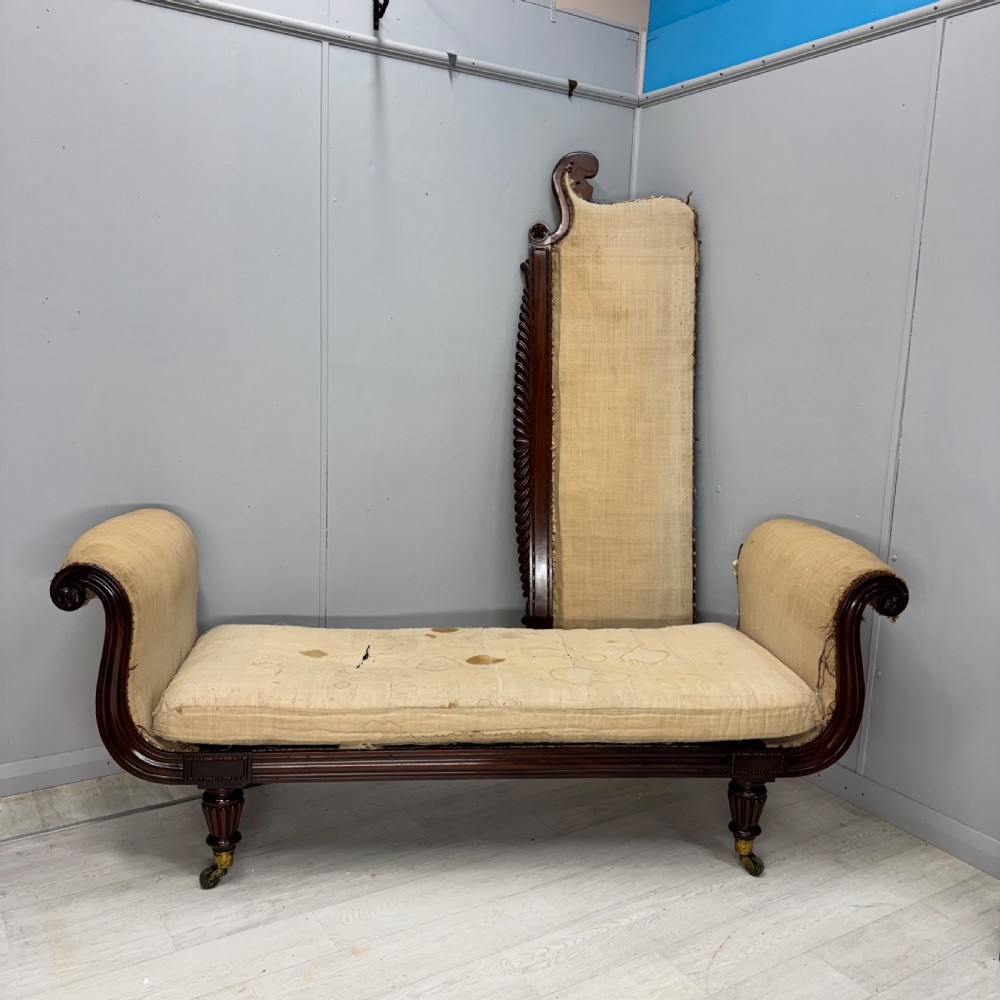 An antique Regency mahogany sofa frame featuring carved scroll arms, fluted turned legs on brass castors, and calico upholstery prepared for recovering. View X