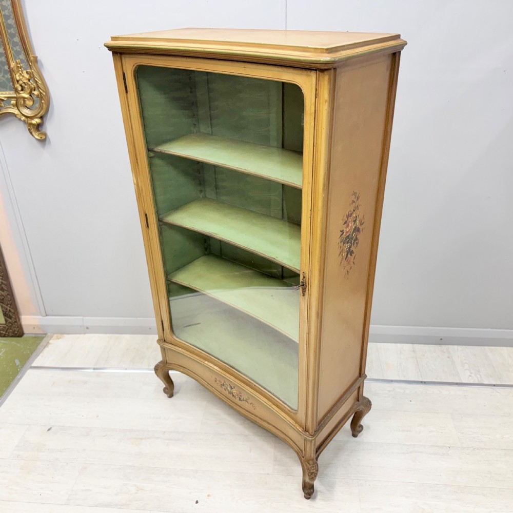 A freestanding cream painted bookcase featuring a single curved glass door, floral painted motifs, cabriole legs, and a sage green interior with adjustable shelves.